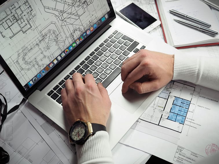 Architect reviewing detailed blueprints and schematics on a laptop with a smartphone nearby.